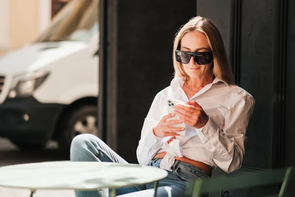 Elegancka biała kobieta siedząca przy stoliku w kawiarni i pisząca smsy na smartfonie, pewna siebie przedsiębiorczyni nosząca okulary przeciwsłoneczne i białą koszulę czekając na spotkanie. — Zdjęcie stockowe