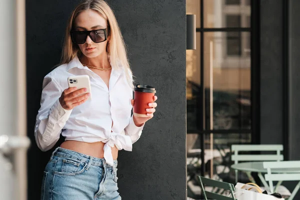 Элегантная женщина, стоящая у стены на террасе кафе, держа в руках напиток, используя смартфон, уверенный в себе предприниматель в солнечных очках в ожидании встречи в кафе. — стоковое фото