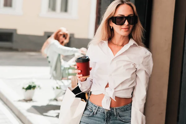 Elegante Frau kommt aus einem Café, hält eine Tasse Kaffee in der Hand und schaut weg, selbstbewusste Unternehmerin trägt Sonnenbrille, Coffee to Go-Konzept. — Stockfoto