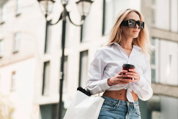Elegancka blondynka trzymająca filiżankę kawy w dłoni i patrząca w dal, pewna siebie przedsiębiorczyni nosząca okulary przeciwsłoneczne i złotą biżuterię Spacerująca ulicą miasta. — Zdjęcie stockowe