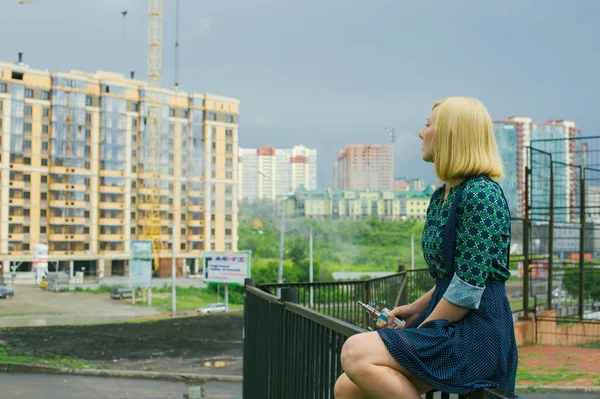 Hermosa joven sentada sobre un fondo de paisaje urbano elektronuuyu fuma un cigarrillo — Foto de Stock