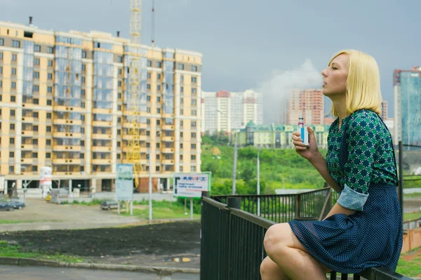 Hermosa joven sentada sobre un fondo de paisaje urbano elektronuuyu fuma un cigarrillo — Foto de Stock