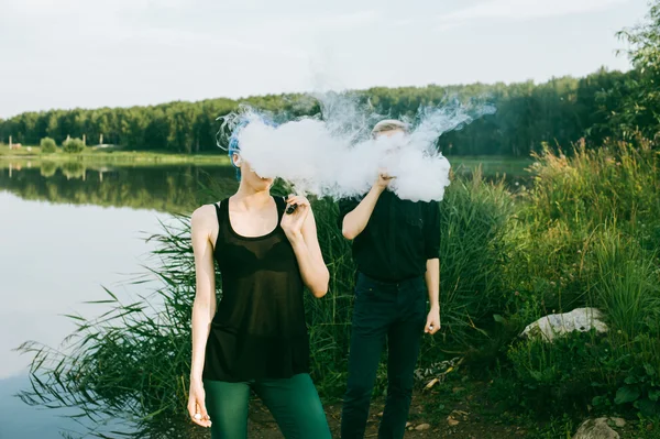 Jovem casal com cabelos tingidos fumar cigarros eletrônicos no lago — Fotografia de Stock