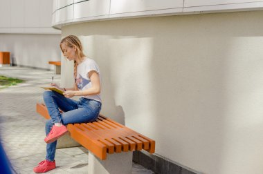 Genç kız öğrenci, dışarıdaki avludaki bankta oturan bir not defterine çizer ve yazar. Çalışmayla açık hava eğlencesini birleştiriyor. Uzun saçlı, beyaz tişörtlü ve kot pantolonlu.