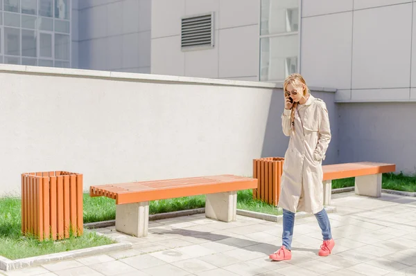 Junges schönes Mädchen mit langen Haaren, die vom Wind angefacht werden, in einem Regenmantel, der auf der Straße mit dem Handy telefoniert. Telefonkommunikation, Liaison, angenehme Gespräche über das Geschäft — Stockfoto