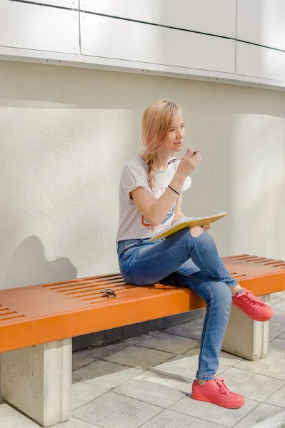 Genç kız öğrenci, dışarıdaki avludaki bankta oturan bir not defterine çizer ve yazar. Çalışmayla açık hava eğlencesini birleştiriyor. Uzun saçlı, beyaz tişörtlü ve kot pantolonlu. — Stok fotoğraf