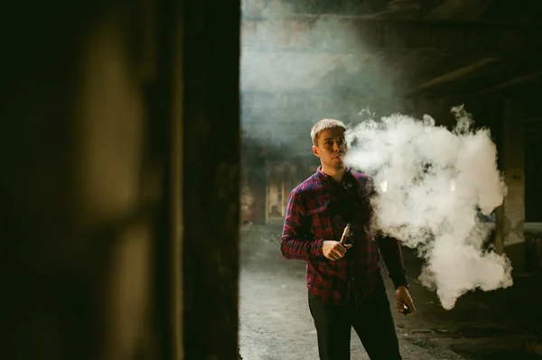 Hombre en una camisa a cuadros y jeans, fuma un cigarrillo electrónico — Foto de Stock
