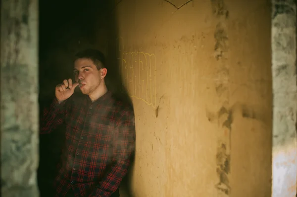 Άνδρας σε ένα καρό πουκάμισο και τζιν, καπνίζει ένα ηλεκτρονικό τσιγάρο, φυσώντας καπνό και ατμός από την εξάτμιση μια μηχανική συσκευή. τοποθεσία εγκαταλειφθεί Ημιτελές κτίριο, τοίχοι από τούβλα, κομψό. — Φωτογραφία Αρχείου