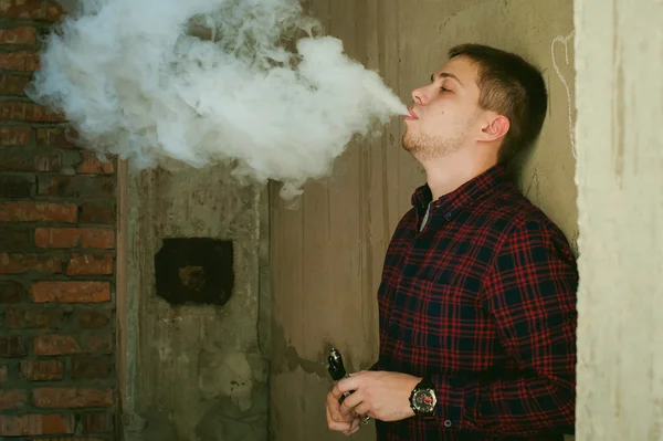 Hombre en una camisa a cuadros y jeans, fuma un cigarrillo electrónico —  Fotos de Stock