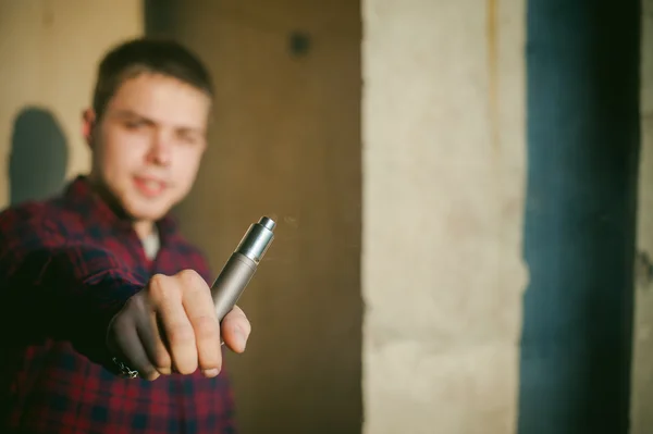 Man in a plaid shirt and jeans, smokes an electronic cigarette, blowing smoke and steam from the vaporization a mechanical device. location Abandoned unfinished building, brick walls, stylish. — Stock Photo, Image