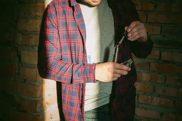 Hombre en una camisa a cuadros y pantalones vaqueros se ejecuta el jugo de vapor cigarrillo electrónico. Tiene un mod mecánico con RDA . — Foto de Stock