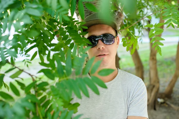 Giovane ragazzo in occhiali da sole fisico standard in posa per strada su uno sfondo di cespugli verdi. Indossa una camicia chiara e un berretto verde scuro. ritratto di un uomo giorno d'estate — Foto Stock
