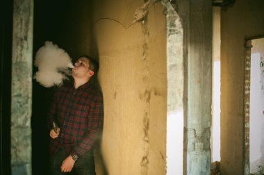 adam bir ekose gömlek ve kot pantolon, duman ve buharlaşma Buhar mekanik bir aygıt üfleme elektronik sigara, Sigara içiyor. bitmemiş bina, tuğla duvarlar, şık konumu terk.