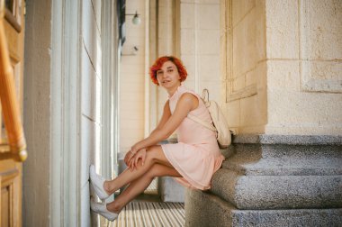 bacaklar yukarı tutan sütunlar arasında oturan beyaz sırt çantası ile bir soluk pembe elbiseli boyalı kırmızı saçlı kadın