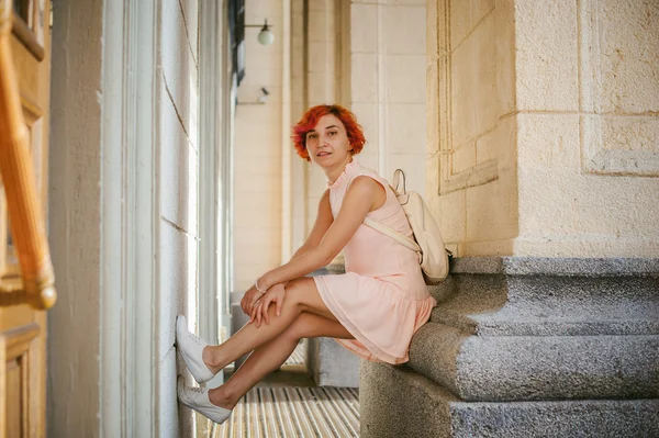 Mulher com cabelos vermelhos tingidos em um vestido rosa pálido com mochila branca, sentado entre colunas segurando as pernas — Fotografia de Stock