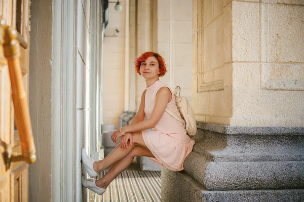 woman with dyed red hair in a pale pink dress with white backpack, sitting between columns holding up the legs