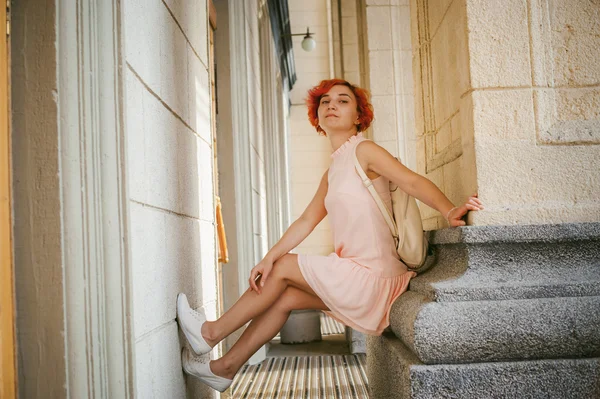 woman with dyed red hair in a pale pink dress with white backpack, sitting between columns holding up the legs