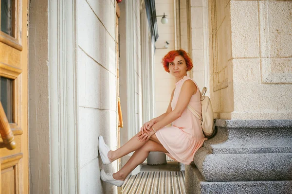 woman with dyed red hair in a pale pink dress with white backpack, sitting between columns holding up the legs