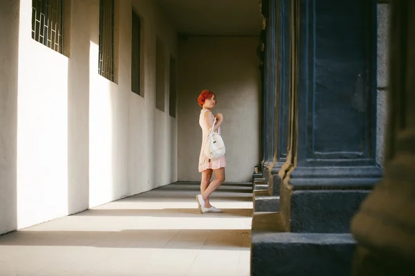 Mädchen in einem blassrosa Kleid mit gefärbten roten Haaren, einen weißen Rucksack in der Hand, wandelt zwischen Licht und Schatten auf den Säulen sonniger Sommertag — Stockfoto