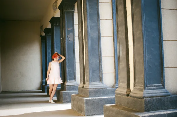 Niña en un vestido de color rosa pálido con el pelo rojo teñido, una mochila blanca en la mano, caminando entre la luz y la sombra en las columnas soleado día de verano —  Fotos de Stock