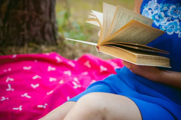 Блондинка в синем платье сидит на красном одеяле на зеленой траве, читает книгу посреди сосновых лесов — стоковое фото