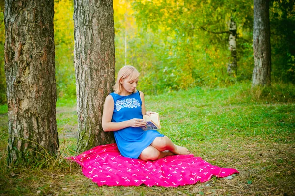 Chica rubia en un vestido azul sentado en una manta roja en la parte superior de la hierba verde, leyendo un libro en medio de árboles de pinos — Foto de Stock