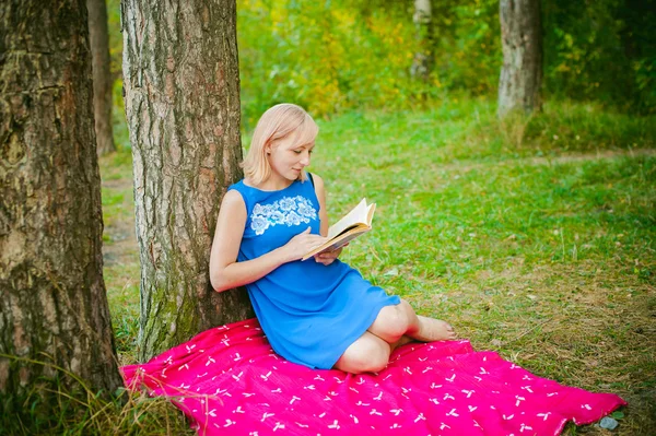 Sarışın kız mavi elbiseli kırmızı battaniye yeşil çimenlerin üstüne oturan bir çam ormanı ağaçlarının ortasında bir kitap okuma — Stok fotoğraf