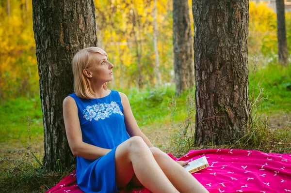 Ragazza bionda con un vestito blu seduta su una coperta rossa in cima all'erba verde, leggendo un libro in mezzo agli alberi di pineta — Foto Stock