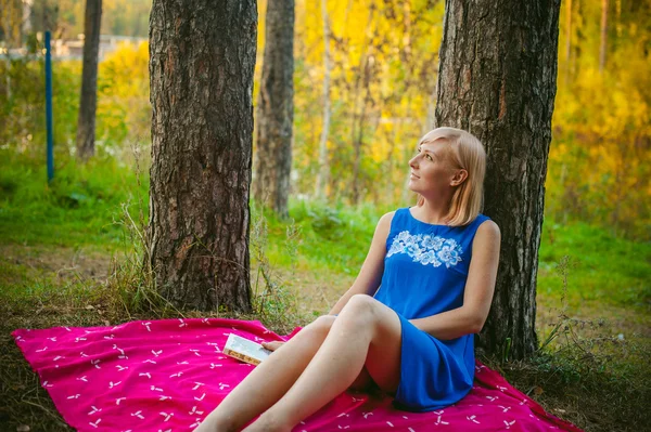 Ragazza bionda con un vestito blu seduta su una coperta rossa in cima all'erba verde, leggendo un libro in mezzo agli alberi di pineta — Foto Stock