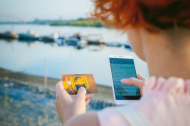 gün batımında, nehir kenarında yürüyüş kırmızı saçlı kız, aygıttan bir banka banka kartı kullanarak online alışveriş için ödeme yapmak için. Online ödeme, banka havaleleri.