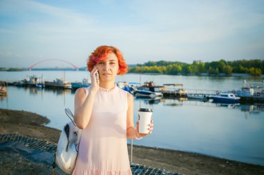Kızıl saçlı ve nehir banka yürüyüş sırt çantası soluk pembe elbiseli kız, telefonda konuşurken ve zemin teknelerin karşı bir karton bardak kahve içme demirlemiş bir sıcak yaz gününde
