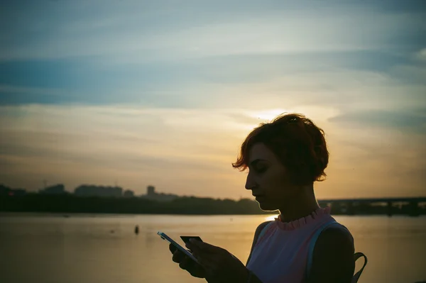 Ragazza con i capelli rossi a piedi lungo il fiume al tramonto, per effettuare pagamenti per gli acquisti online dal dispositivo utilizzando una carta di debito bancario. pagamenti online, bonifici bancari . — Foto Stock