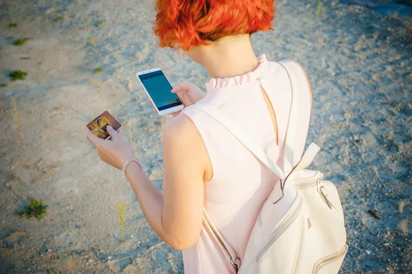 Menina com cabelos vermelhos andando pelo rio ao pôr do sol, para fazer pagamentos para compras on-line a partir do seu dispositivo usando um cartão de débito bancário. pagamentos em linha, transferências bancárias . — Fotografia de Stock