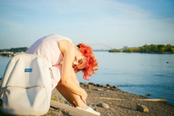 Mädchen in blassrosa Kleid mit roten Haaren und Rucksack am Ufer des Flusses sitzend am sandigen Ufer — Stockfoto