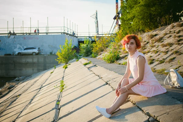 Niña en vestido rosa pálido con el pelo rojo y la mochila caminando a lo largo de la orilla del río, sentado en la orilla arenosa — Foto de Stock