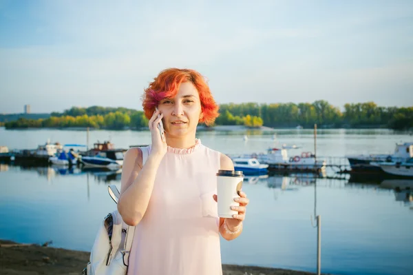Menina em vestido rosa pálido com cabelo vermelho e mochila andando ao longo da margem do rio, falando ao telefone e beber café de uma xícara de papelão, contra o pano de fundo de barcos ancorados em um dia quente de verão — Fotografia de Stock