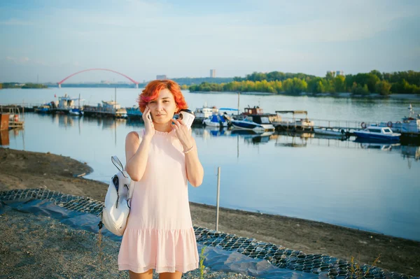 Menina em vestido rosa pálido com cabelo vermelho e mochila andando ao longo da margem do rio, falando ao telefone e beber café de uma xícara de papelão, contra o pano de fundo de barcos ancorados em um dia quente de verão — Fotografia de Stock