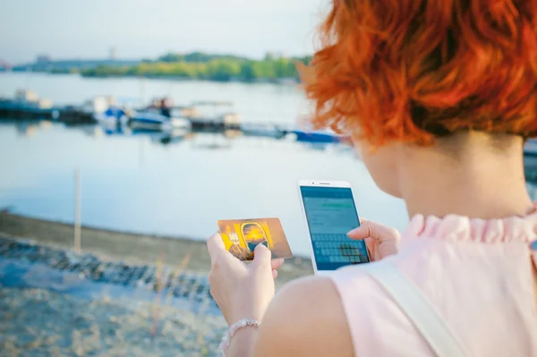 Gadis dengan rambut merah berjalan di tepi sungai saat matahari terbenam, untuk melakukan pembayaran untuk pembelian online dari perangkat Anda menggunakan kartu debit bank. pembayaran online, transfer bank . Stok Gambar