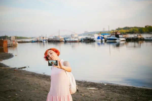 Dziewczyna w blado różowa sukienka z rude włosy i plecak spacer wzdłuż brzegu rzeki, zdjęcia z siebie na ich telefon fotoradar, na tle łodzie zacumowane na ciepły, letni dzień — Zdjęcie stockowe