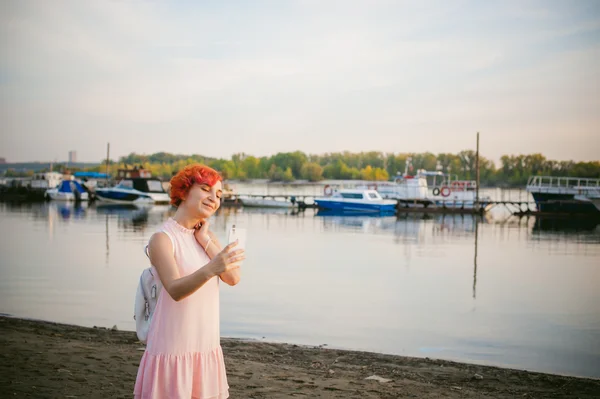 Девушка в бледно-розовом платье с рыжими волосами и рюкзак прогуливаясь вдоль берега реки, фотографии себя на мобильном телефоне камеры, на фоне лодок пришвартованых в теплый летний день — стоковое фото
