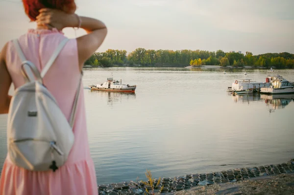 Girl in pale pink dress with red hair and backpack walking along river bank, pictures of themselves on their mobile camera phone, against backdrop of boats moored on a warm summer day — Stock Photo, Image