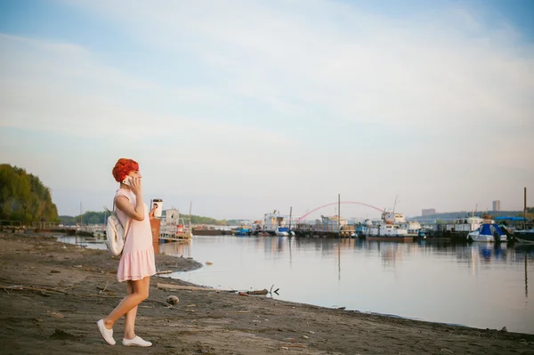 Девушка в бледно-розовом платье с рыжими волосами и рюкзаком идет вдоль берега реки, разговаривает по телефону и пьет кофе из картонной чашки, на фоне лодок пришвартованых в теплый летний день — стоковое фото
