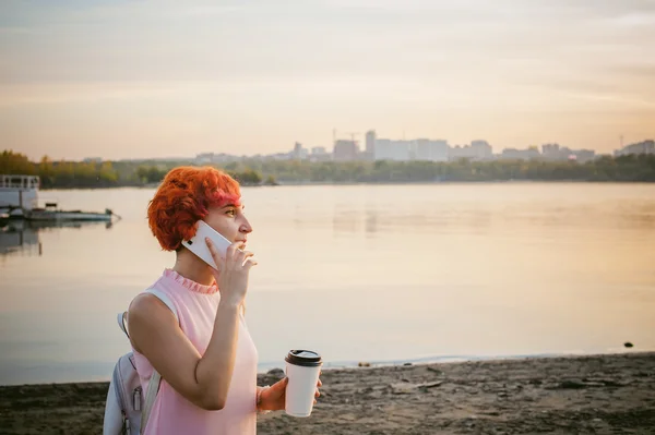 Ragazza in abito rosa pallido con capelli rossi e zaino passeggiando lungo la riva del fiume, parlando al telefono e bevendo caffè da una tazza di cartone, sullo sfondo di barche ormeggiate in una calda giornata estiva — Foto Stock