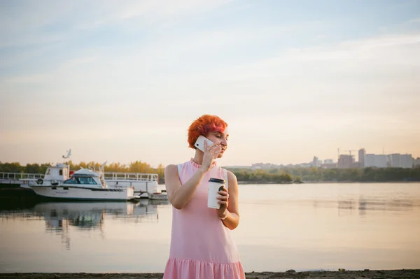 Ragazza in abito rosa pallido con capelli rossi e zaino passeggiando lungo la riva del fiume, parlando al telefono e bevendo caffè da una tazza di cartone, sullo sfondo di barche ormeggiate in una calda giornata estiva — Foto Stock