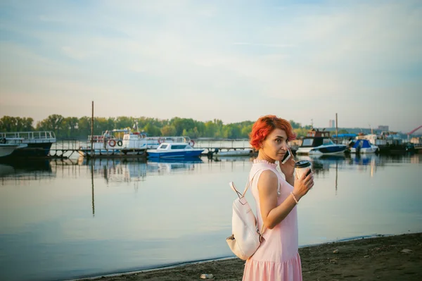 Fille en robe rose pâle avec cheveux roux et sac à dos marchant le long de la rive de la rivière, parlant au téléphone et buvant du café dans une tasse en carton, sur fond de bateaux amarrés par une chaude journée d'été — Photo