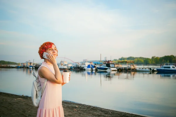 Dívka v světle růžové šaty s červenými vlasy a batohem pěšky podél břehu řeky, mluvil po telefonu a pití kávy z hrnečku kartonové pozadí lodě zakotvené na teplý letní den — Stock fotografie