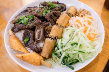 Vienamese Pho vermicelli, ızgara sığır eti, kızarmış çıtır börek, balık çubuğu, yeşil soğan, doğranmış marul, salatalık, havuç ve turp dilimleri beyaz bir tabakta servis edilir.