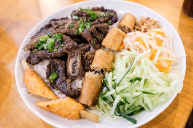 Vienamese Pho vermicelli, ızgara sığır eti, kızarmış çıtır börek, balık çubuğu, yeşil soğan, doğranmış marul, salatalık, havuç ve turp dilimleri beyaz bir tabakta servis edilir.