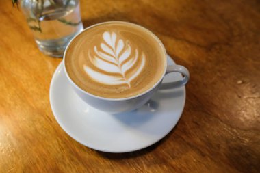 Kafe latte kahvenin yatay fotoğrafını beyaz fincanda bir yaprak tablosu, içinde çiçek olan bir cam şişe ile kapat.