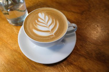 Kafe latte kahvenin yatay fotoğrafını beyaz fincanda bir yaprak tablosu, içinde çiçek olan bir cam şişe ile kapat.